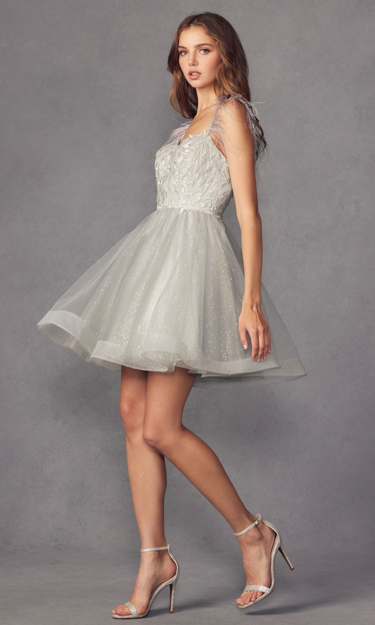 A woman wearing the Short Babydoll Homecoming Dress with Feathers 881 and silver high-heeled sandals stands against a plain gray background.